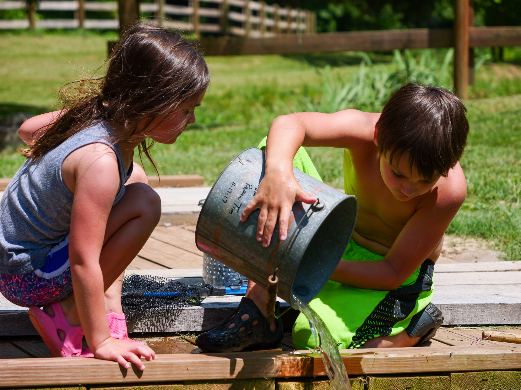 Two kids playing with a bucket of water on a wooden deck on a sunny summer day in Hawkins County, Tennessee.